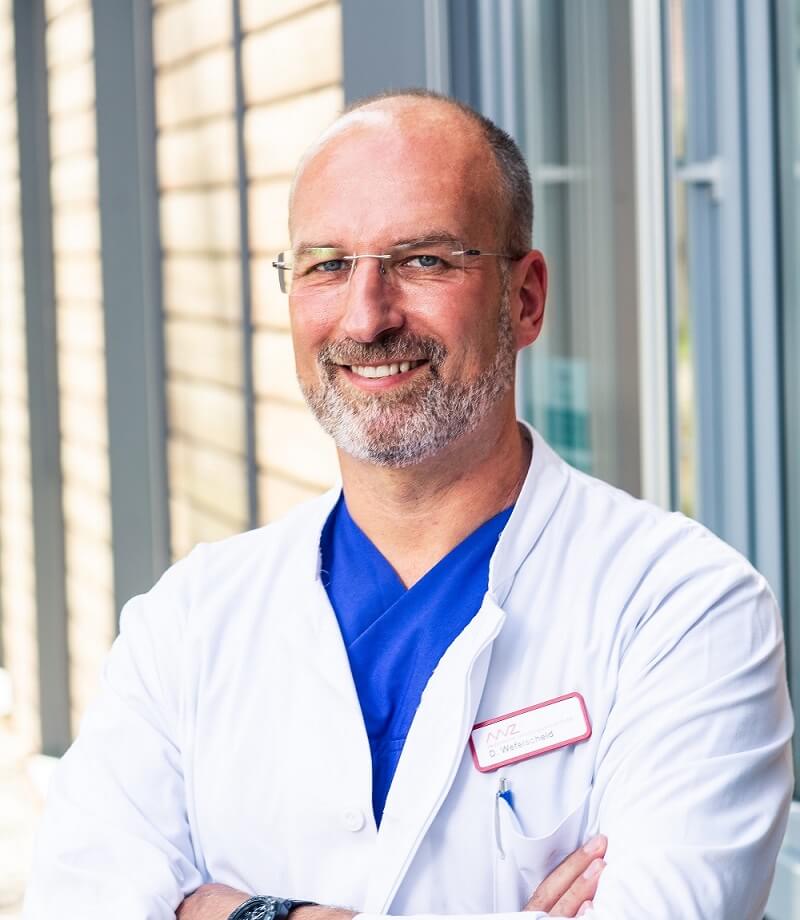 Sie sehen einen lächelnden Mann mittleren Alters mit Halbglatze und weiß-gräulichem Bart. Er trägt eine eckige rahmenlose Brille und einen weißen Arztkittel über einem blauen Kassak.